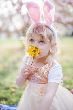Küçük sevimli kız Manolya yakın çim üzerinde oturuyor. Bir Paskalya tavşanı gibi giyinmiş bir kız bir çiçek ve bir yumurta tutar.