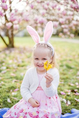Küçük sevimli kız Manolya yakın çim üzerinde oturuyor. Bir Paskalya tavşanı gibi giyinmiş bir kız bir çiçek ve bir yumurta tutar.