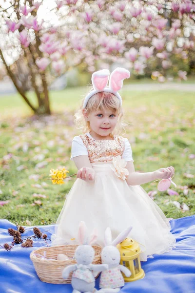 Küçük sevimli kız Manolya yakın çim üzerinde oturuyor. Bir Paskalya tavşanı gibi giyinmiş bir kız bir çiçek ve bir yumurta tutar. — Stok fotoğraf