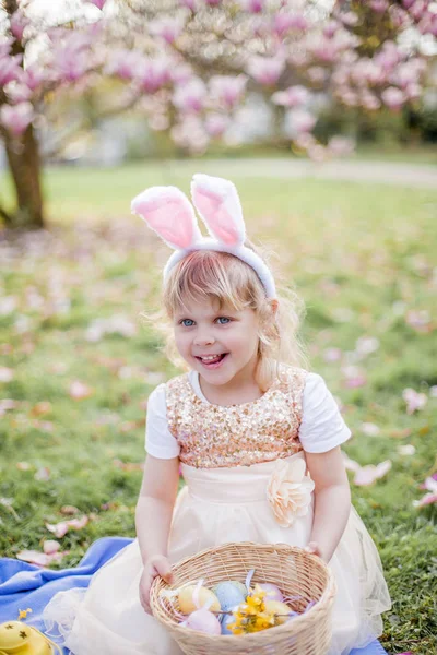Mała śliczna dziewczyna siedzi na trawie w pobliżu Magnolia. Dziewczyna ubrana jako króliczka Wielkanocna posiada kwiat i jajko. — Zdjęcie stockowe