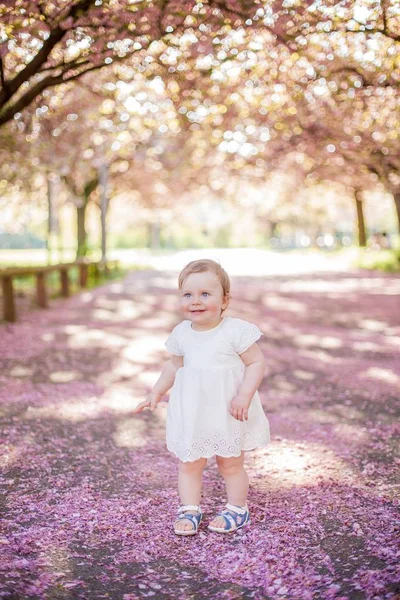 Niña en un vestido blanco corre en el parque con flores de color rosa. Primavera . —  Fotos de Stock