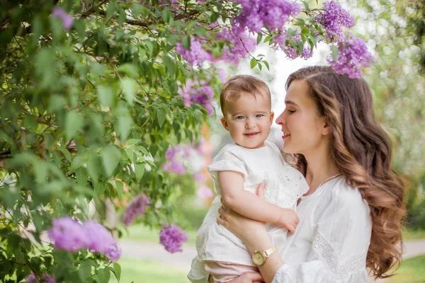 Hermosa madre joven y pequeña hija linda son felices cerca de la lila. Retrato de mamá y su hija. Primavera . —  Fotos de Stock