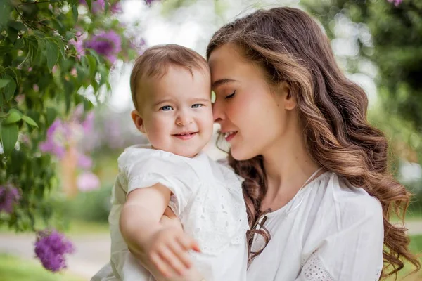 Bella giovane madre e piccola figlia carina sono felici vicino al lilla. Ritratto di mamma e figlia. Primavera . — Foto Stock