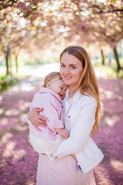 Güzel genç anne ve küçük sevimli kızı çiçekli bir parkta yürürken mutluyuz. Anne ve kızının portresi. Kiraz çiçekleri.