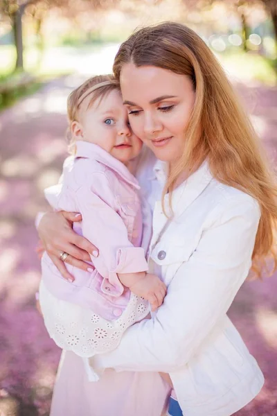 Beautiful young mother and little cute daughter are happy walking in a flowered park. Portrait of mom and daughter. Cherry blossoms. — Stock Photo, Image