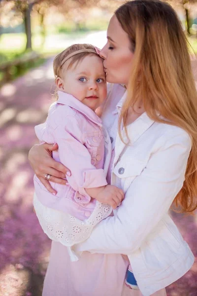 Beautiful young mother and little cute daughter are happy walking in a flowered park. Portrait of mom and daughter. Cherry blossoms. — Stock Photo, Image