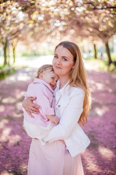 Krásná mladá matka a malá roztomilá dceruška jsou šťastni, když chodí do květného parku. Portrét matky a dcery. Třešňové květy. — Stock fotografie