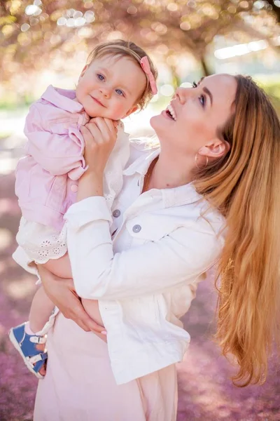 Gyönyörű fiatal anya és a kis aranyos lánya boldog séta egy virágos parkban. Portré az anya és lánya. Cseresznye virágok. — Stock Fotó