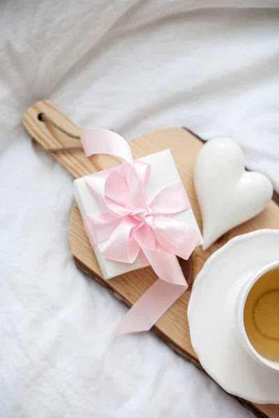 Caneca branca com chá, caixa de presente com fita na cama. Pequeno-almoço na cama. Aconchegante. Xadrez rosa. Algodão . — Fotografia de Stock