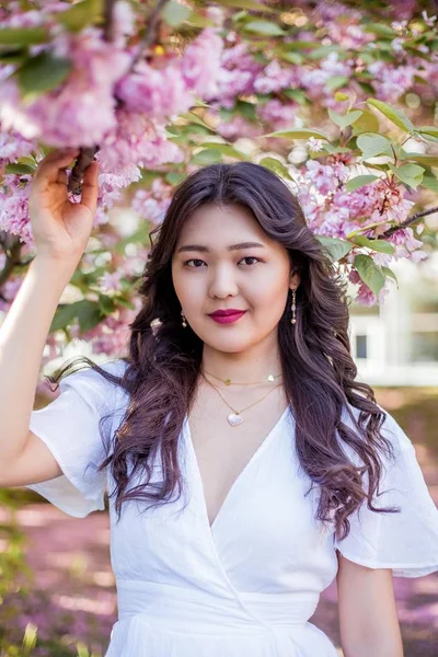 Uma jovem mulher asiática bonita em um vestido branco caminha em um parque florido. Sakura. Plantas em flor . — Fotografia de Stock