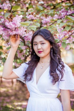 Beyaz elbiseli genç ve güzel bir Asyalı kadın çiçekli bir parkta yürüyor. Sakura. Çiçek açan ağaçlar.