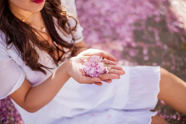 Close-up. Ženské ruce drží plátky růžových květů. Mladá krásná asijská žena v bílých šatech prochází v květém parku. — Stock fotografie