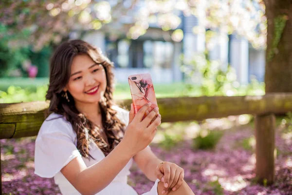 Młoda piękna Azjatycka Kobieta w białej sukni spacery w ukwieconym parku. Młoda piękna Azjatycka kobieta robi selfie na telefon. Sakura. Kwitnące drzewa. — Zdjęcie stockowe