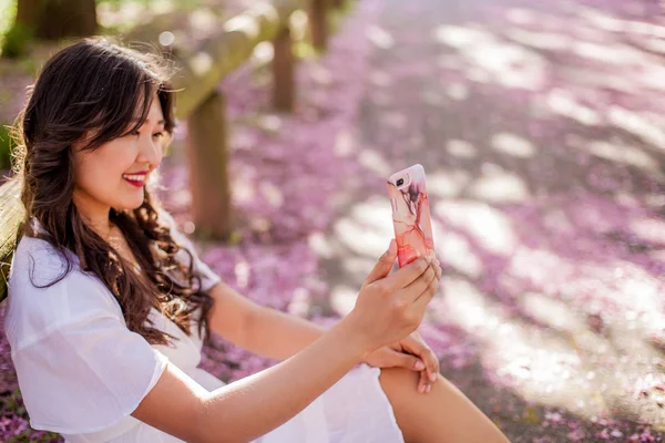Młoda piękna Azjatycka Kobieta w białej sukni spacery w ukwieconym parku. Młoda piękna Azjatycka kobieta robi selfie na telefon. Sakura. Kwitnące drzewa. — Zdjęcie stockowe