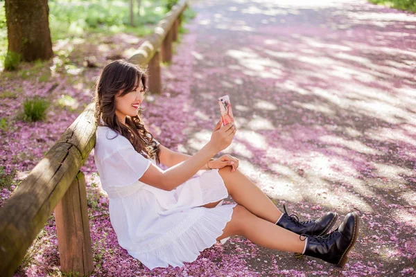 Młoda piękna Azjatycka Kobieta w białej sukni spacery w ukwieconym parku. Młoda piękna Azjatycka kobieta robi selfie na telefon. Sakura. Kwitnące drzewa. — Zdjęcie stockowe