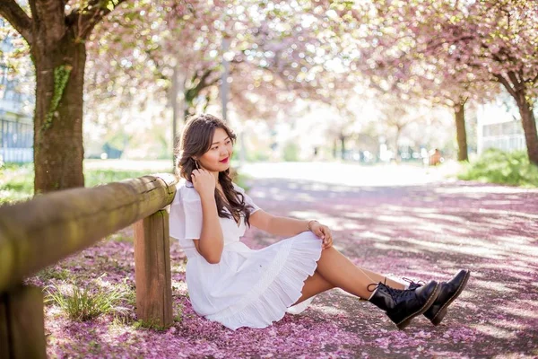 Uma jovem mulher asiática bonita em um vestido branco caminha em um parque florido. Sakura. Plantas em flor . — Fotografia de Stock