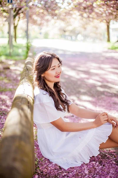 Uma jovem mulher asiática bonita em um vestido branco caminha em um parque florido. Sakura. Plantas em flor . — Fotografia de Stock