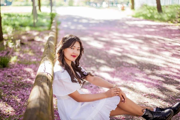 Uma jovem mulher asiática bonita em um vestido branco caminha em um parque florido. Sakura. Plantas em flor . — Fotografia de Stock