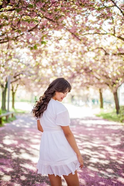 白いドレスを着た若い美しいアジア人女性が花の咲く公園を歩く。さくら。咲く木々. — ストック写真