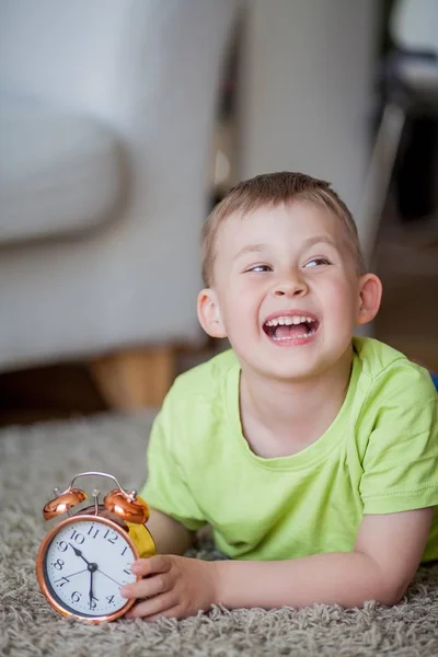 Söt liten pojke på 4 år gammal ligger på mattan och håller en väckarklocka i hans händer. Morgon. Uppvaknande. — Stockfoto