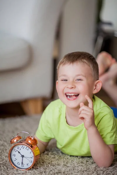 Schattige kleine jongen van 4 jaar oud ligt op het tapijt en in de buurt van de wekker. Ochtend. Ontwaken. — Stockfoto