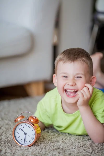 Schattige kleine jongen van 4 jaar oud ligt op het tapijt en in de buurt van de wekker. Ochtend. Ontwaken. — Stockfoto