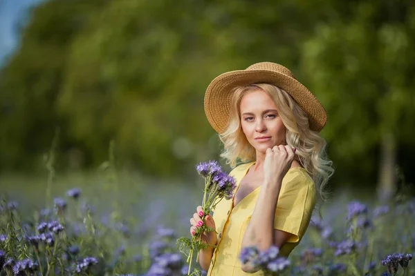 Die junge schöne Blondine mit Hut geht durch ein Feld lila Blumen. Sommer. — Stockfoto