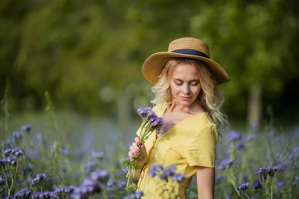 Die junge schöne Blondine mit Hut geht durch ein Feld lila Blumen. Sommer. — Stockfoto