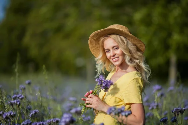 Die junge schöne Blondine mit Hut geht durch ein Feld lila Blumen. Sommer. — Stockfoto