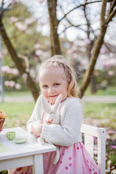 Dos niñas rubias lindas de 3 años están jugando en el parque cerca de una magnolia floreciente. Beber té. Semana Santa . — Foto de Stock