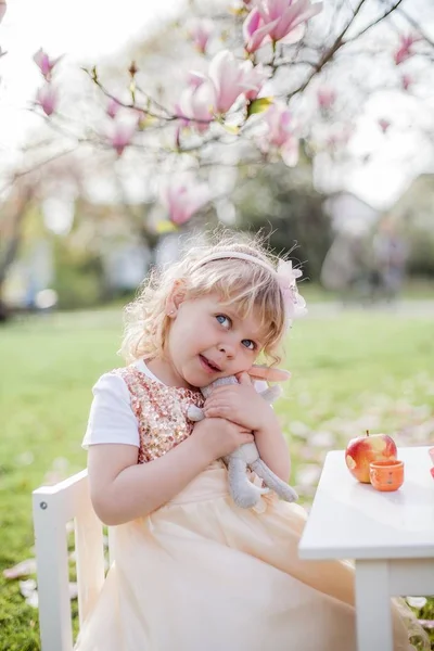 Dos niñas rubias lindas de 3 años están jugando en el parque cerca de una magnolia floreciente. Beber té. Semana Santa . —  Fotos de Stock