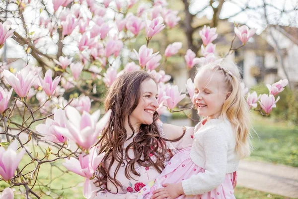 Bela jovem mãe e filhinha perto de uma magnólia florescente. Primavera. Flores cor de rosa . — Fotografia de Stock