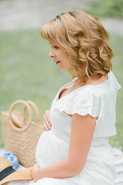 Ung vacker gravid kvinna i vit klänning sitter på ängen och äter jordgubbar. Picknick. Moderskap. — Stockfoto