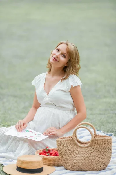 Wanita muda hamil dengan gaun putih duduk di padang rumput dan makan stroberi. Piknik. Keibuan . — Stok Foto
