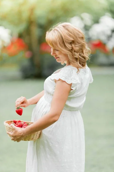 Wanita muda hamil cantik dengan gaun putih berjalan melalui taman dan makan stroberi. Piknik. Keibuan . — Stok Foto