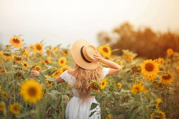 Bela jovem encaracolado em um campo de girassol segurando um chapéu de vime. Retrato de uma jovem ao sol. Verão . — Fotografia de Stock