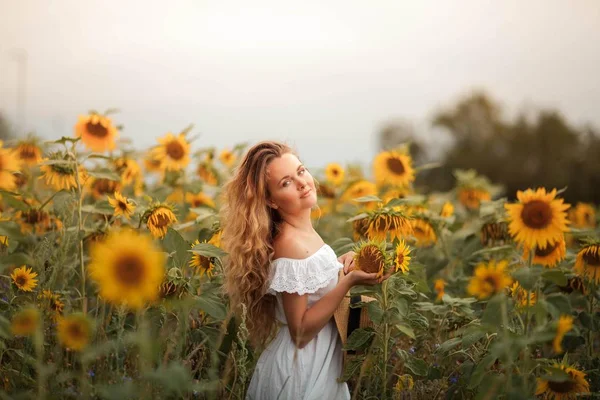 Bela jovem encaracolado em um campo de girassol segurando um chapéu de vime. Retrato de uma jovem ao sol. Verão . — Fotografia de Stock