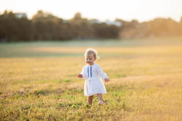 Una niña rizada de ojos azules con un vestido blanco camina por el campo. Retrato de una niña al sol. Puesta de sol . —  Fotos de Stock