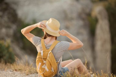 Sırt çantası olan şapkalı güzel kızıl saçlı genç kadın dağlardaki bir uçurumda oturuyor. Turizm.