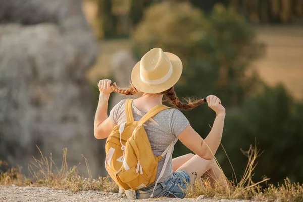 Jovem ruiva bonita em um chapéu com uma mochila senta-se em um penhasco nas montanhas. Turismo . — Fotografia de Stock