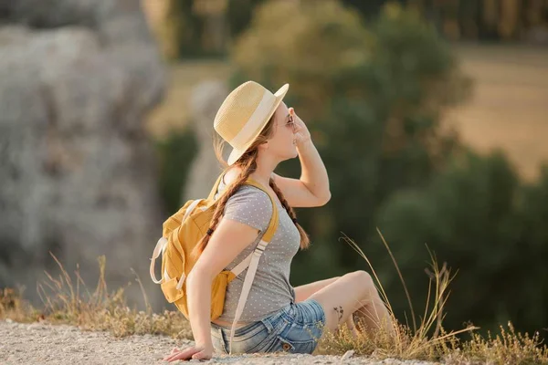 Jovem ruiva bonita em um chapéu com uma mochila senta-se em um penhasco nas montanhas. Turismo . — Fotografia de Stock