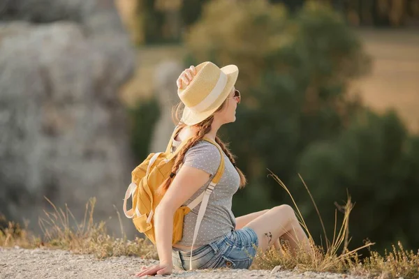 Jovem ruiva bonita em um chapéu com uma mochila senta-se em um penhasco nas montanhas. Turismo . — Fotografia de Stock