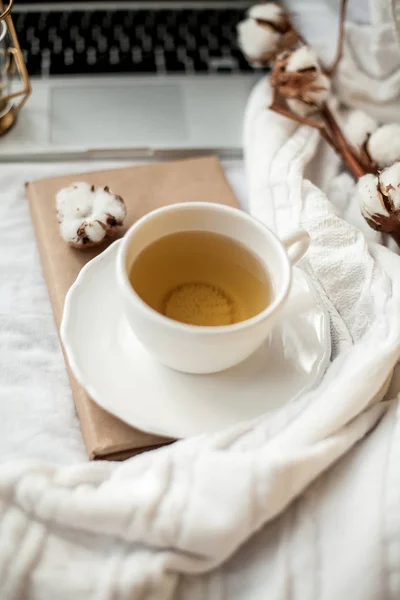 A white mug with tea, cotton, a laptop, a plaid, a candle and a book stand on the bed. Breakfast in bed. Cozy. Autumn. Winter. — Stock Photo, Image