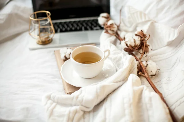 Uma caneca branca com chá, algodão, um laptop, uma xadrez, uma vela e um suporte de livro na cama. Pequeno-almoço na cama. Aconchegante. Outono. Inverno . — Fotografia de Stock