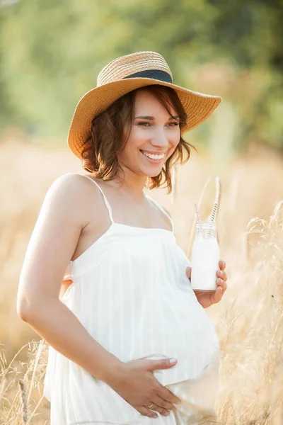 Wanita muda hamil yang cantik di ladang gandum meminum susu dari botol kaca. Potret seorang wanita hamil muda di bawah sinar matahari. Musim panas . — Stok Foto
