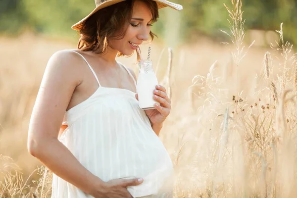 Vacker ung gravid kvinna i ett vetefält dricker mjölk från en glas flaska. Porträtt av en ung gravid kvinna i solen. Sommaren. — Stockfoto