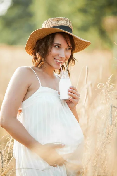 Beautiful young pregnant woman in a wheat field drinks milk from a glass bottle. Portrait of a young pregnant woman in the sun. Summer. — Stock Photo, Image