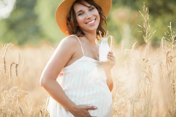 Wanita muda hamil yang cantik di ladang gandum meminum susu dari botol kaca. Potret seorang wanita hamil muda di bawah sinar matahari. Musim panas . — Stok Foto