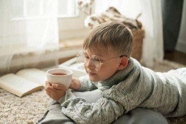 Örme kazak giyen güzel bir çocuk pencerenin yanında bir fincan sıcak çayla kitap okuyor. Rahat. Sonbahar.