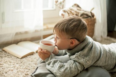 Örme kazak giyen güzel bir çocuk pencerenin yanında bir fincan sıcak çayla kitap okuyor. Rahat. Sonbahar.
